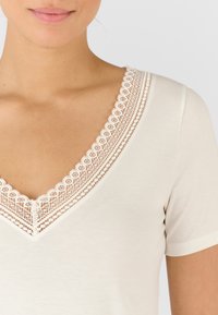T-shirt color crema con scollo a V, caratterizzato da un delicato bordo in pizzo lungo il collo. Maniche corte e texture morbida e liscia.