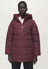 Burgundská prošívaná bunda s kapucí, s předním zapínáním na zip, vodorovným šitím a širokými rukávy. Nošena přes světle růžovou košili.