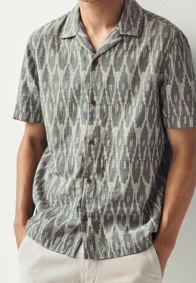 Homme portant une chemise à manches courtes boutonnée avec un motif géométrique abstrait gris et beige et un pantalon beige, mains dans les poches.