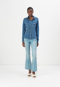 Camicia di jeans blu scuro con maniche lunghe, colletto a punta e due tasche sul petto. Abbinata a jeans a palazzo blu chiaro e sandali bianchi.