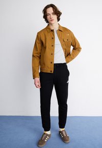 Bruine canvas jas, grijze t-shirt, zwarte joggingsbroek met logo, bruine sneakers. Model poseert tegen een effen achtergrond met blauwe vloer.