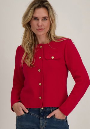 Vrouw met lang blond haar, gekleed in een rode jas met gouden knopen en blauwe spijkerbroek, staand met handen in de zakken.