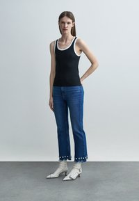 Woman standing wearing a black tank top with white trim, blue jeans with floral-embellished frayed hems, white socks, and white heels.