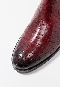 Bottine en cuir synthétique rouge imitation crocodile avec un bout pointu, texture brillante et une semelle noire lisse avec des détails subtils.