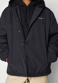 Person i en svart huva Carhartt-jacka över en svart hoodie med händerna i fickorna och beigea byxor synliga nedanför.