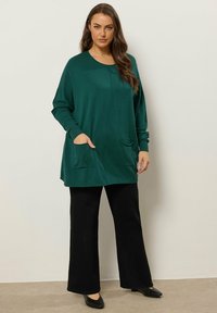 Cardigan lungo color verde scuro realizzato in morbido tessuto a maglia, con scollatura rotonda, maniche lunghe e due tasche anteriori. Abbinato a pantaloni neri.
