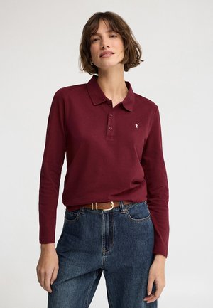 Mujer vistiendo una camisa polo de manga larga color granate metida en unos vaqueros azules de talle alto con un cinturón marrón sobre un fondo liso.