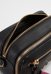 Černá syntetická kožená crossbody taška se zlatými zipy, několika přihrádkami, texturovaným povrchem a akcentním popruhem v růžové barvě.