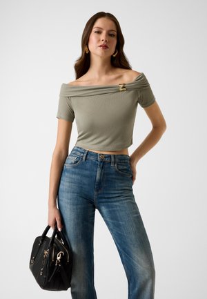 Femme portant un crop top vert olive à épaules dénudées avec une épingle dorée, un jean bleu, tenant un sac à main noir sur un fond uni.