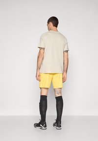 Lys beige t-skjorte, gule shorts, svarte knestrømper og svarte sko. Tatoveringer på armene; enkel tekstur. Minimalistisk design uten synlige mønstre.