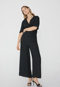 Mono negro de pierna ancha con escote en V, tejido texturizado, mangas cortas y cintura ceñida. Combinado con sandalias de tacón alto negras con tiras.