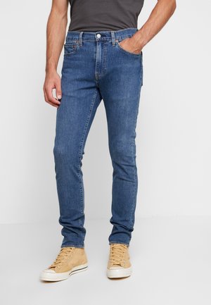Homme portant un jean slim bleu, une main dans la poche, associé à des baskets montantes beige et un t-shirt sombre.