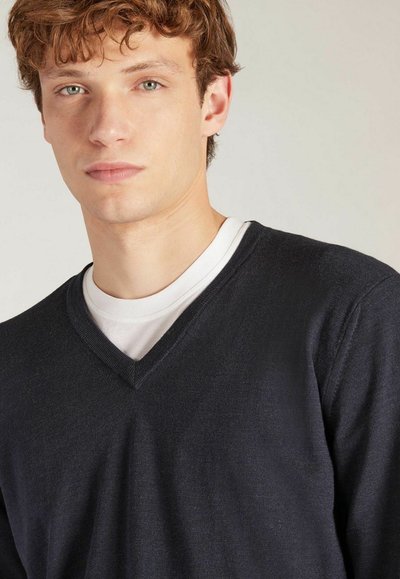 Marineblauer V-Ausschnitt-Pullover aus einem weichen, strukturierten Stoff, über ein weißes Rundhalsausschnitt-Shirt getragen. Schlichtes, sauberes Design ohne Muster.