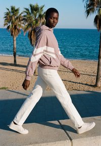 Persoană tânără cu pantaloni albi, windbreaker lila deschis cu dungi albe și adidași albi, plimbându-se pe o plajă cu palmieri și ocean în fundal.