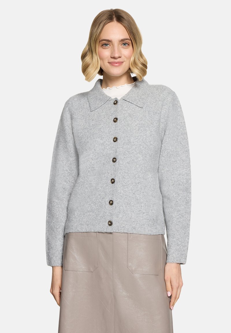 Femme aux cheveux blonds mi-longs portant un cardigan gris clair boutonné sur une jupe beige avec poches à l'avant, devant un fond uni.
