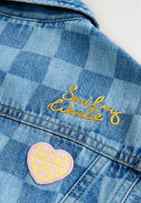 Jeansjacke mit einem hellblauen Karomuster, verziert mit gelb gesticktem Text "Smiley World" und einem herzförmigen Patch in Pink mit Schachbrettmuster.