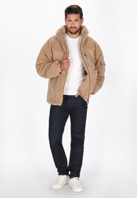 Tan fleecejakke med tekstureret overflade, beige paneler, justerbar hætte, lynlåslommer, parret med mørke jeans og hvide sneakers.