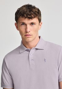Polo de color púrpura claro hecho de algodón, con un cuello clásico, una tapeta con tres botones y un pequeño logo en el lado izquierdo del pecho.