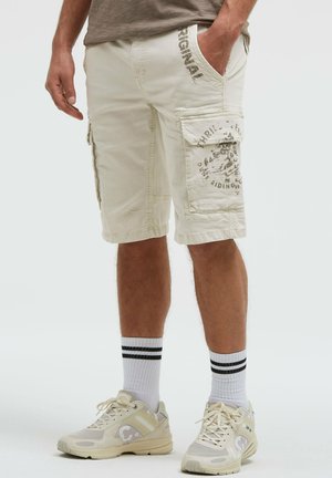 Mann trägt beige Cargo-Shorts mit aufgedrucktem Text, weiße Sportsocken mit schwarzen Streifen und cremefarbene Sneakers, steht vor einem einfarbigen Hintergrund.