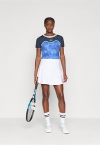 Marineblå og blå tie-dye trenings-T-skjorte med grå kant, sammen med en hvit skjørt, holder en svart og blå tennisrackett.