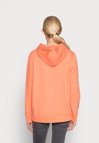 Henkilö, jolla on vaaleat hiukset poninhännällä, pukeutuneena yksinkertaiseen oranssiin huppariin ja tummanharmaisiin housuihin, kääntynyt pois valkoista taustaa vasten.