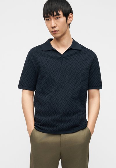 Άνδρας που φοράει υφασμένη κοντομάνικη μπλούζα πόλο σε σκούρο ναυτικό μπλε με γιακά τύπου κάμπινγκ και χακί παντελόνι, στέκεται με τα χέρια στις τσέπες.