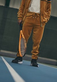 Oranžna teniška raketa, ki jo drži oseba v zlatem velur trenirki z črnimi detajli in Nike supergami, na modrem teniškem igrišču.