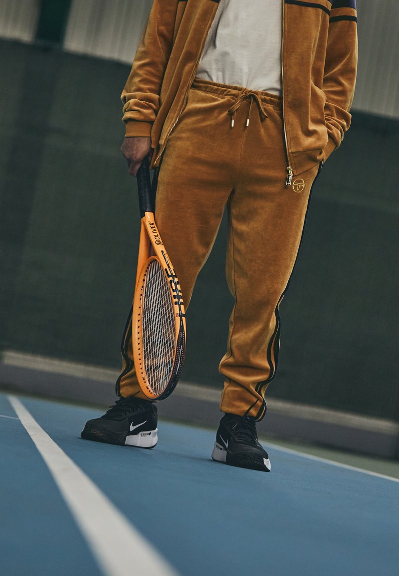 Oranžna teniška raketa, ki jo drži oseba v zlatem velur trenirki z črnimi detajli in Nike supergami, na modrem teniškem igrišču.