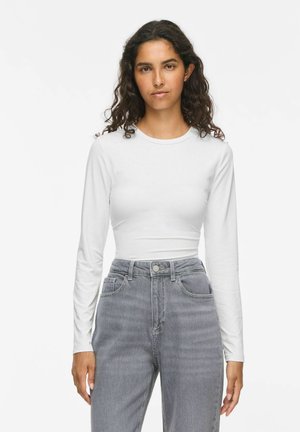 Femme aux longs cheveux bouclés portant une chemise blanche ajustée à manches longues et un jean gris taille haute, debout devant un fond blanc uni.
