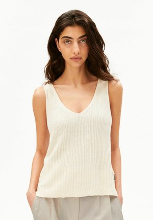 Femme aux longs cheveux foncés portant un haut en maille sans manches à col en V de couleur crème et un pantalon beige clair les mains dans les poches.