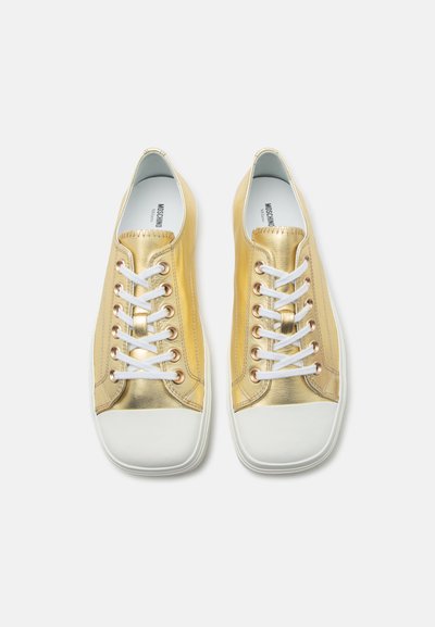 Ζευγάρι μεταλλικά χρυσά αθλητικά παπούτσια χαμηλού πάτου με λευκές λαστιχένιες μύτες, λευκά κορδόνια και λευκές σόλες σε απλό φόντο.