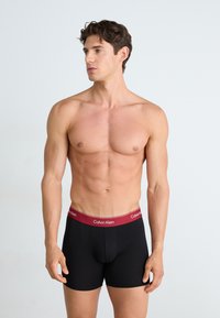 Zwarte boxershorts met een bordeauxrode tailleband met het Calvin Klein-logo, gedragen door een model dat naar rechts kijkt, en een slim fit ontwerp toont.