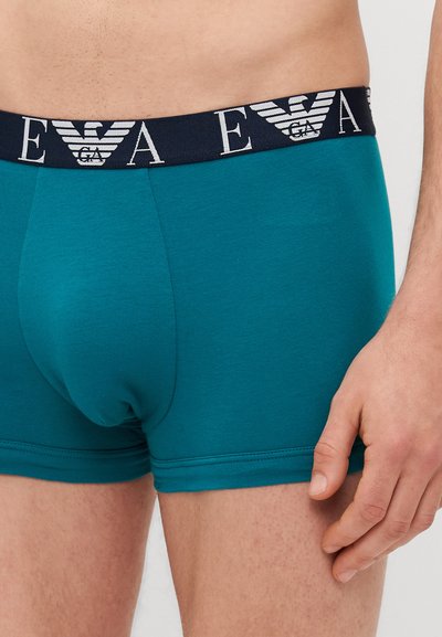 Tyrkysové boxerky vyrobené z mäkkej tkaniny s tmavomodrým elastickým pásom s bielym logom. Strih je priliehavý s viditeľnými švami.