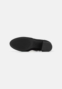 Chaussure noire en daim avec une semelle texturée, présentant un talon noir lisse. La base a des rainures horizontales pour une meilleure adhérence.