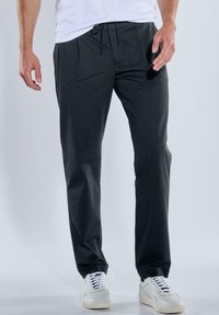 Dunkelgraue Hose mit Kordelzugbund, gefälteltem Vorderteil und schmal zulaufendem Bein. Aus glattem Stoff gefertigt, kombiniert mit weißen Sneakers.