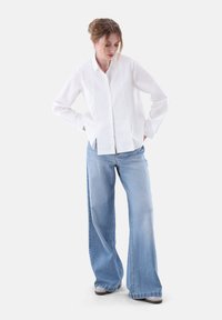 Camicia bianca con colletto e maniche lunghe, abbinata a jeans blu chiaro a gamba larga. Tessuto morbido, vestibilità casual, senza motivi visibili.
