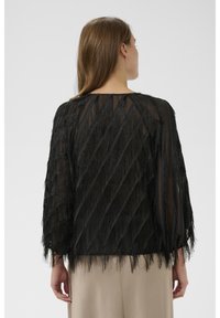 Top negru transparent cu detaliu texturat, franjuri diagonali. Croială lejeră cu mâneci lungi, decolteu rotund și drapaj elegant.