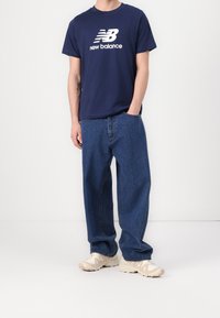 Navy blue puuvillapaita, jossa on valkoinen "New Balance" -logo, yhdistettynä löysiin farkkuihin ja beigeihin urheilukengiin.