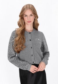 Cárdigan gris con mangas adornadas con lentejuelas, escote redondo, cierre de botones en el frente y un sutil bolsillo en el pecho. Lleva puesto sobre una falda negra.