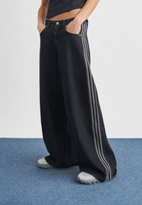 Person wearing wide-leg black jeans with three gray side stripes, hands in pockets, paired with white sneakers on blue carpet.