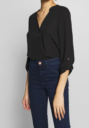 Vrouw draagt een zwarte blouse met lange mouwen die zijn opgerold en donkerblauwe high-waisted jeans tegen een effen achtergrond.