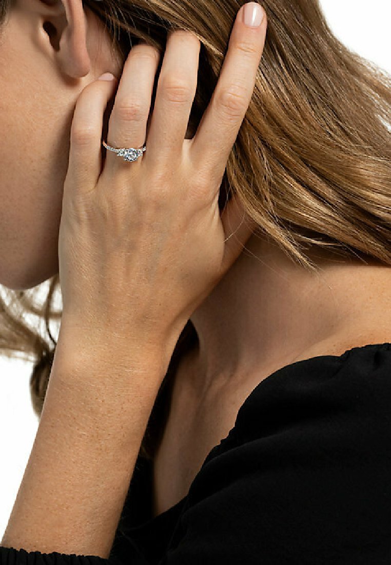 Bague de fiançailles en diamant avec une pierre centrale, un anneau en argent et des détails complexes. Une main repose près du visage, mettant en valeur l'éclat de la bague.