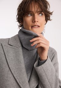 Osoba z krótkimi brązowymi włosami, ubrana w szary wełniany płaszcz i sweter typu golf, dotyka kołnierza i spogląda w bok zamyślona.