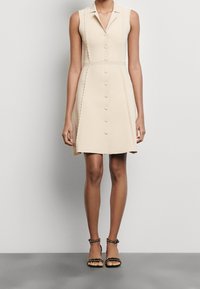 Vestito beige senza maniche con colletto, apertura frontale con bottoni e dettagli ricamati sui lati. Indossato con sandali neri con borchie decorative.