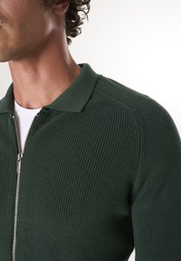 Zelený pletený svetr na zip s ribovanou texturou, klasickým límcem a stříbrným zipem s červeným akcentem.