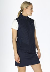 Gilet blu navy con pannello anteriore trapuntato, design aderente e dettagli riflettenti, indossato sopra una camicia bianca a maniche corte e una gonna blu navy.