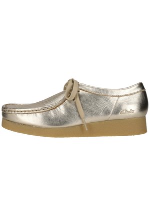 Skinnende gylden læder mokkasinsko med snørebånd, beige crepesål og rundet tå, mærket med Clarks-logo på hælsiden.