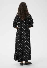 Fekete maxi ruha fehér pöttyökkel, rövid ujjú és egyenes szabású. Fekete lapos cipővel viselve, hátulnézetből.