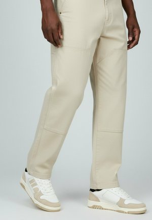 Pantaloni beige con cuciture dettagliate e cuciture laterali, abbinati a sneakers bianche e beige con un design testurizzato e chiusura con lacci.