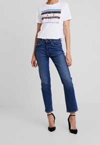 Kvinna i vit grafisk t-shirt, blå högmidjade raka jeans och svarta högklackade sandals med ankelrem mot en enfärgad bakgrund.